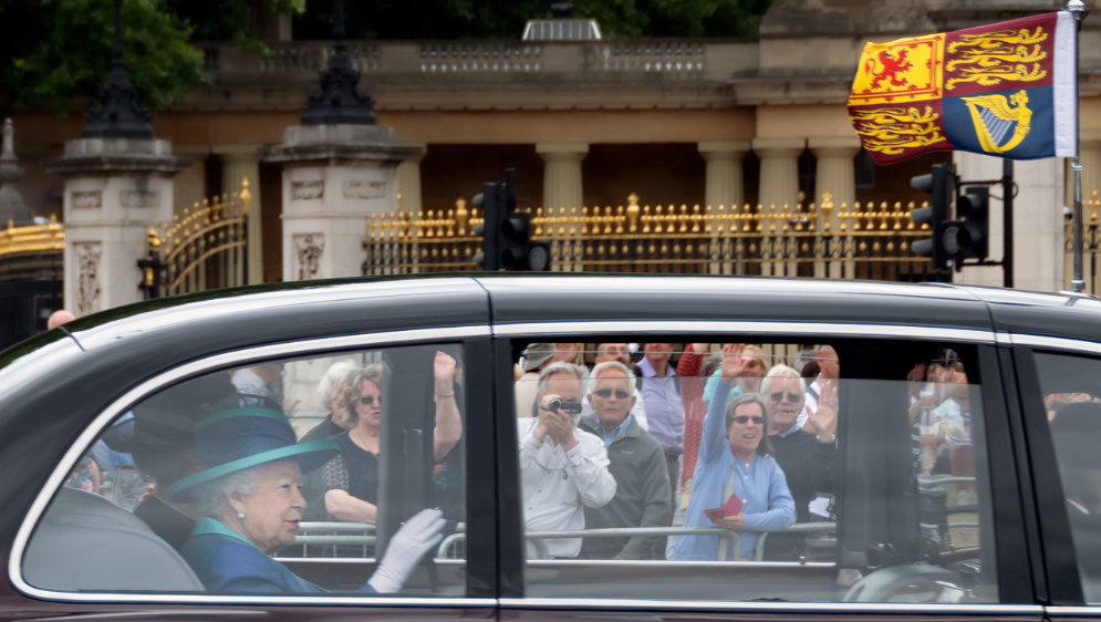 The Queen waving from a car with the Royal Standard flying.